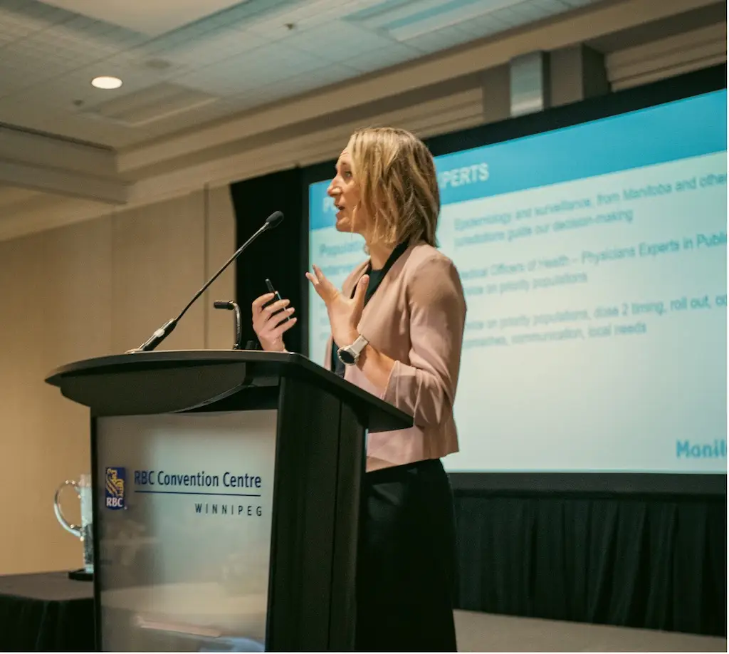 A photo of Dr. Joss Reimer standing before a microphone. A glass jug of water and cups are on a table beside her.