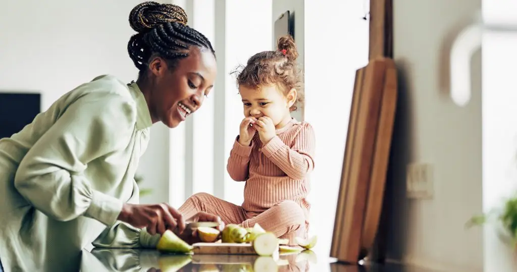 Smiling young African mom and her adorable little girl enjoying a healthy fruit snack together in their kitchen at home