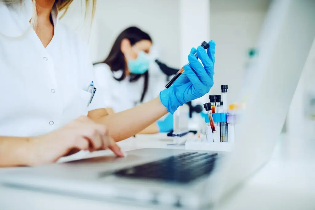 Female lab assistant using laptop for data entry and in other hand holding test tube with blood sample while sitting in laboratory.