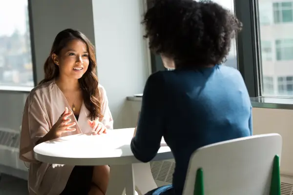 Two women sitting at a white table. The woman closest to us as he back to us. She is in a navy shirt. The woman facing us is an Asian woman with long highlighted curled hair. She is smirking while explaining something. She has earring and rings on.