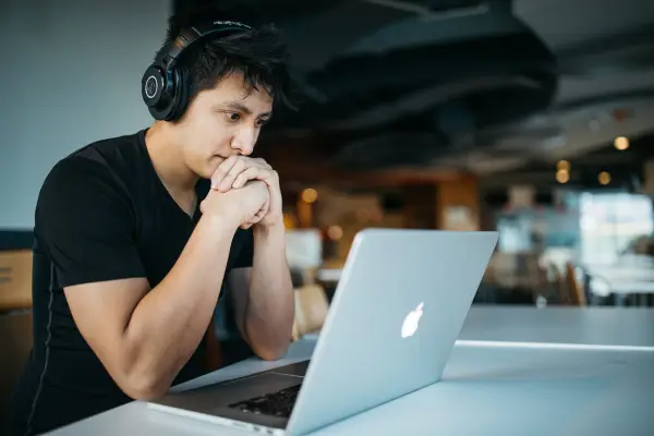 photo of Asian man in a black shirt with over the ear black headphones. He is sitting at a table looking intently at a MacBook. the background is blurred and can be assumed he is in an empty coffee shop.